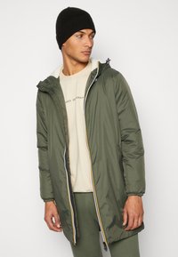 Olive green puffer jacket with a hood, white inner lining, and contrasting zipper details. Worn over a beige t-shirt and green pants.