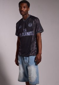 Camiseta negra con el texto "LA FRATERNITÉ", patrón intrincado y mangas cortas, combinada con shorts de denim azul claro desgastados.