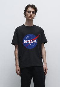 Černé bavlněné tričko s kulatým modrým logem NASA, bílé texty a červený oblouk. Model nosí tmavé džíny.