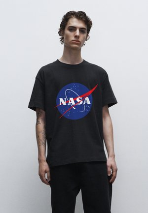 Schwarzes Baumwoll-T-Shirt mit einem kreisförmigen blauen NASA-Logo, weißem Text und einem roten Schwung. Das Modell trägt dunkle Jeans.