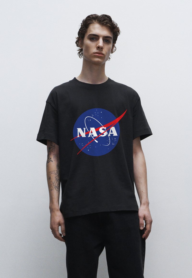 Černé bavlněné tričko s kulatým modrým logem NASA, bílé texty a červený oblouk. Model nosí tmavé džíny.