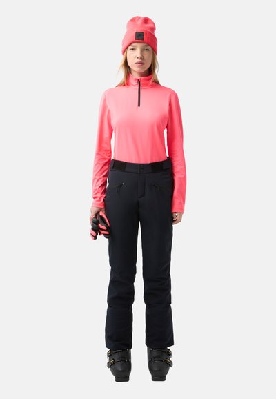 Leuchtend pinkfarbener Longsleeve mit einem Viertelreißverschluss, kombiniert mit schwarzen wasserdichten Hosen mit Reißverschlusstaschen und einem verstärkten Bund. Schwarze Skischuhe.