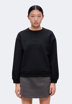 Kvinde med mørkt hår sat tilbage iført en sort sweatshirt med præget logo og en grå nederdel mod en ensartet lys baggrund.
