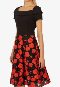 Schwarzes Kleid mit einem strukturierten, schulterfreien Oberteil und einem voluminösen Rock, der mit lebhaften roten Blumenstickereien verziert ist.