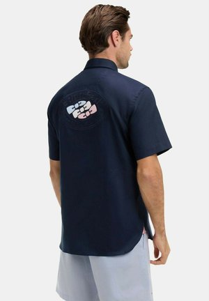 Homme portant une chemise à manches courtes bleu marine avec un graphique aux couleurs pastel dans le dos, associée à un short gris clair, debout devant un fond blanc.