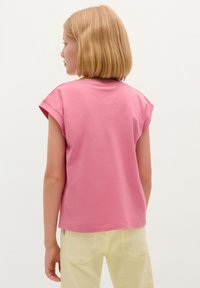 Roze katoenen T-shirt met korte mouwen en een losse pasvorm, gezien vanaf de achterkant, gecombineerd met lichtgele broek. Gladde textuur.