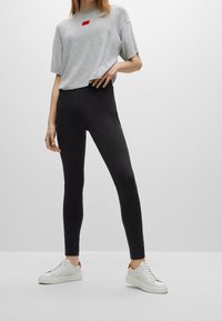 T-shirt gris à manches courtes avec un écusson logo rouge, associé à un legging noir et des baskets blanches avec des accents rouges. Design simple et ajusté.