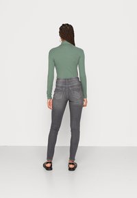 Jeans cinzentos ajustados com dois bolsos traseiros, combinados com uma blusa verde de mangas longas. O tecido tem uma textura suave, e os jeans têm um design clássico.