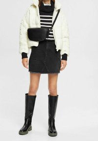 Weiße Puffjacke über einem schwarz-weiß gestreiften Pullover, kombiniert mit einem schwarzen Denim-Rock, knielangen schwarzen Stiefeln und einer schwarzen Umhängetasche.