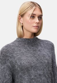 Szary, puszysty sweter z dzianiny o wysokim kołnierzu, z fakturowanym wykończeniem. Modelka nosi małe złote kolczyki w kształcie kółek. Skóra wydaje się gładka i naturalna.