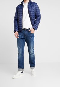 Marineblau gesteppte Jacke mit Stehkragen, seitlichen Reißverschlusstaschen, kombiniert mit blauen Jeans und weißen Sneakers.