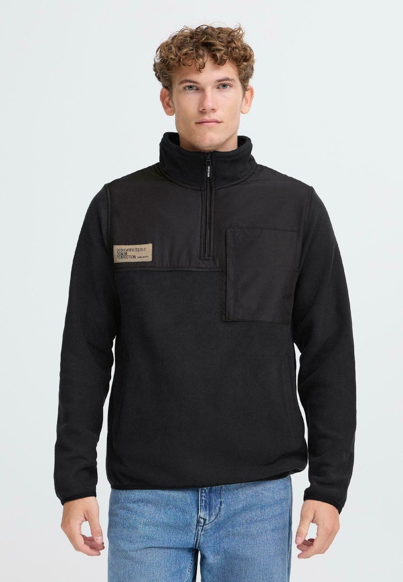 Sudadera de forro polar negra con cuello alto, media cremallera, bolsillo en el pecho, tejido texturizado y un parche de logo en la parte superior izquierda.