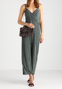 Mono verde texturizado con escote en V profundo y tirantes finos, combinado con un bolso negro bordado con flores. Modelo femenina con sandalias.