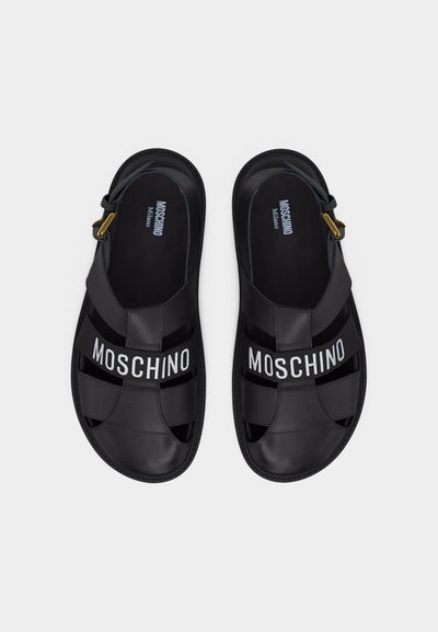 Ζευγάρι μαύρων δερμάτινων σανδαλιών Moschino με κομμένες λουρίδες και λευκή λογότυπη επιγραφή στην πάνω πλευρά, σε λευκό φόντο, φωτογραφημένα από ψηλά.