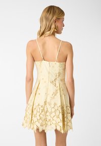 Femme aux cheveux blonds portant une robe jaune en dentelle sans manches avec des bretelles spaghetti, vue de dos sur fond blanc.