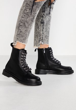 Osoba nosząca czarne skórzane buty wojskowe z metalowymi oczkami oraz szare skrócone jeansy z efektem acid-wash, zamkami błyskawicznymi i postrzępionymi nogawkami.