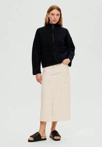 Mulher vestindo um casaco preto com fecho, saia midi creme com fenda frontal e sandálias de plataforma pretas com biqueira aberta, em pé com uma mão no bolso.