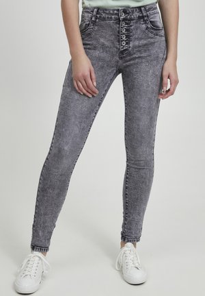 Eng anliegende Jeans in verwaschenem schwarzem Denim mit hoher Taille, ausgestattet mit einem Knopfverschluss und einem Fünf-Taschen-Design. Kombiniert mit weißen Sneakers.