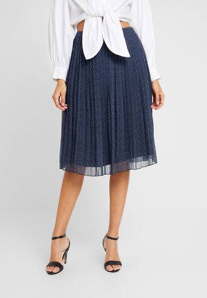 Femme portant une jupe midi plissée bleu marine à petits pois blancs, un chemisier blanc noué et des sandales noires à talons hauts.