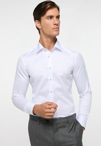 Weißes Langarmhemd mit klassischem Kragen, Einzelknopf-Manschetten und subtiler Sheen, kombiniert mit grauen Hosen in taillierter Passform.