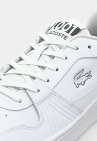 Gros plan sur une basket blanche Lacoste avec des lacets, le texte noir "Lacoste", le numéro "1001" sur la languette, et le logo du crocodile sur le côté.