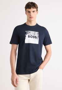 T-shirt di cotone blu navy con grafica quadrata recante la parola "BOSS" in bold nero e un design a pennellate. Maniche corte.