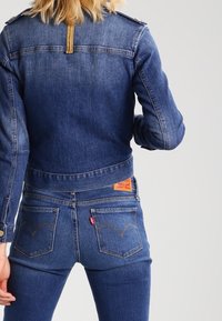 Sininen farkkutakki olkatukin kanssa, jossa on lyhennetty malli ja oranssi brändilogo takana. Yhdistetty samanlaisten farkkujen kanssa.