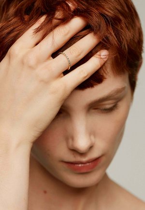 Anillo de oro con una fila de pequeñas piedras preciosas, llevado en una mano que reposa sobre un cabello corto y rojo; el tono de piel es claro con sutiles contornos.
