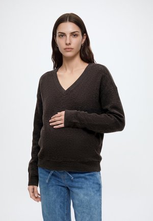 Jeune femme enceinte aux longs cheveux foncés portant un pull marron foncé à col en V et un jean bleu, tenant son ventre d'une main.