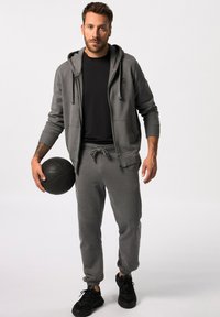 Grijze hoodie met rits en trekkoord, zwart t-shirt, grijze joggingbroek en zwarte basketbalschoenen; houdt een zwarte basketbal vast.