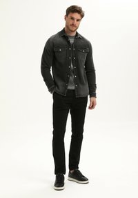 Zwarte denimjack met twee borstzakken over een grijze trui, gecombineerd met zwarte slim-fit broek en zwarte sneakers. Soepel, casual ontwerp.
