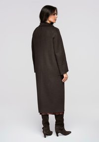 Abrigo largo de color marrón oscuro hecho de mezcla de lana, con un ajuste relajado, mangas anchas y cuello con muesca, combinado con botas marrones hasta la rodilla.