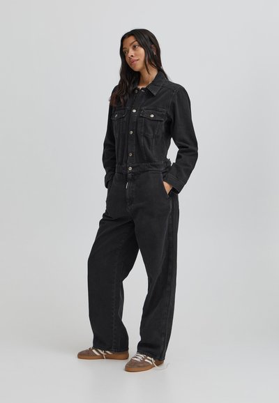 Melnā denima kombinezons ar pieguļošu korpusu, garām piedurknēm un brīviem svārkiem. Iespējas priekšējās kabatas un pogu aizdare. Tekstūrēts materiāls.