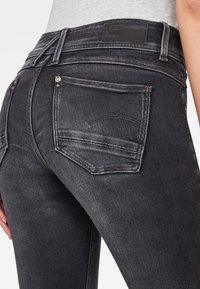 Jeans de mezclilla negra con corte ajustado, que cuentan con dos bolsillos traseros, detalles de costura sutiles y un parche de cuero negro en la cintura.