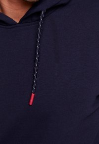 Granatowa bluza z kapturem o miękkiej fakturze, z plecionym sznurkiem w czerwonych akcentach i kontrastowym wzorem na sznurku.