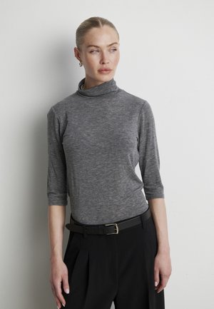 Femme aux cheveux blonds portant un col roulé gris et un pantalon noir, debout contre un mur blanc, regardant vers la droite.