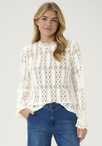 Blusa in pizzo bianco con motivi intricati e maniche lunghe, dotata di polsini arricciati e scollatura rotonda. Abbinata a jeans blu.