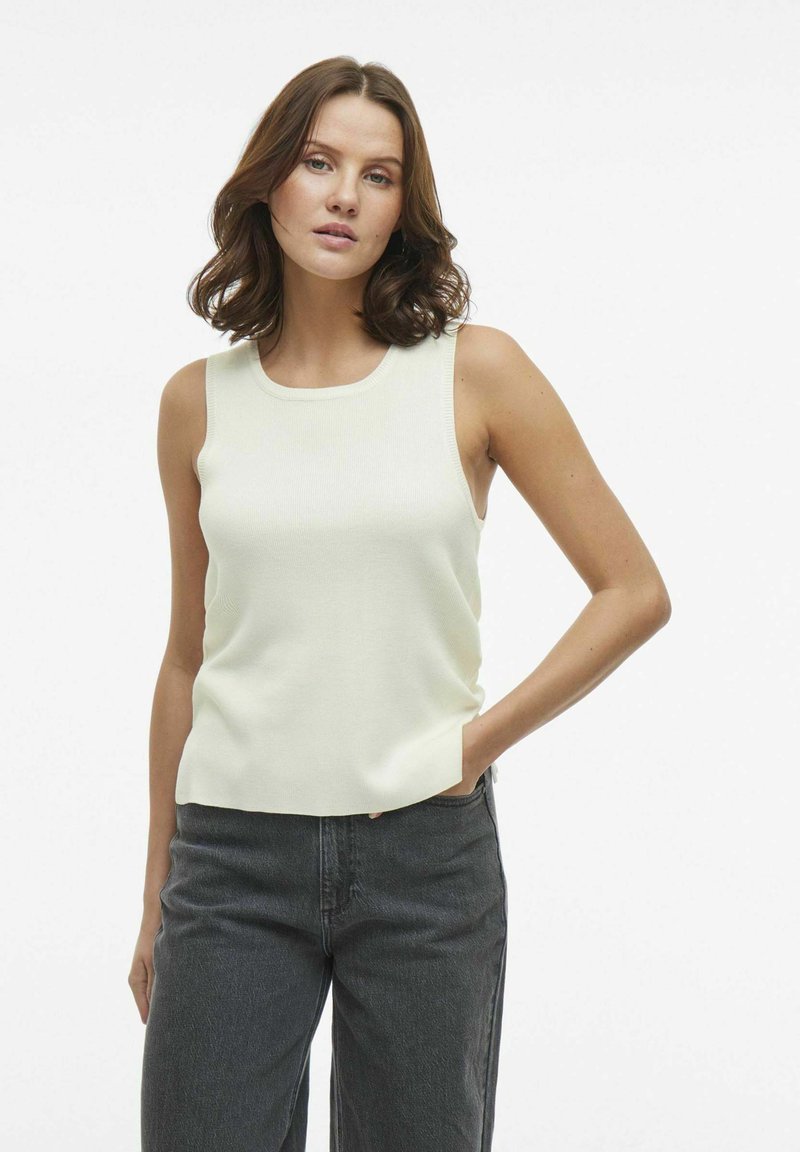 Top sin mangas de color beige claro con escote redondo, textura acanalada y corte relajado, combinado con jeans de talle alto en gris oscuro.