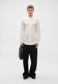 Λευκό πουκάμισο με μακριά μανίκια και όρθιο γιακά, συνδυασμένο με σκούρο τζιν παντελόνι και μαύρη τσάντα χειρός. Ομαλή υφή και εφαρμοστή σχεδίαση.