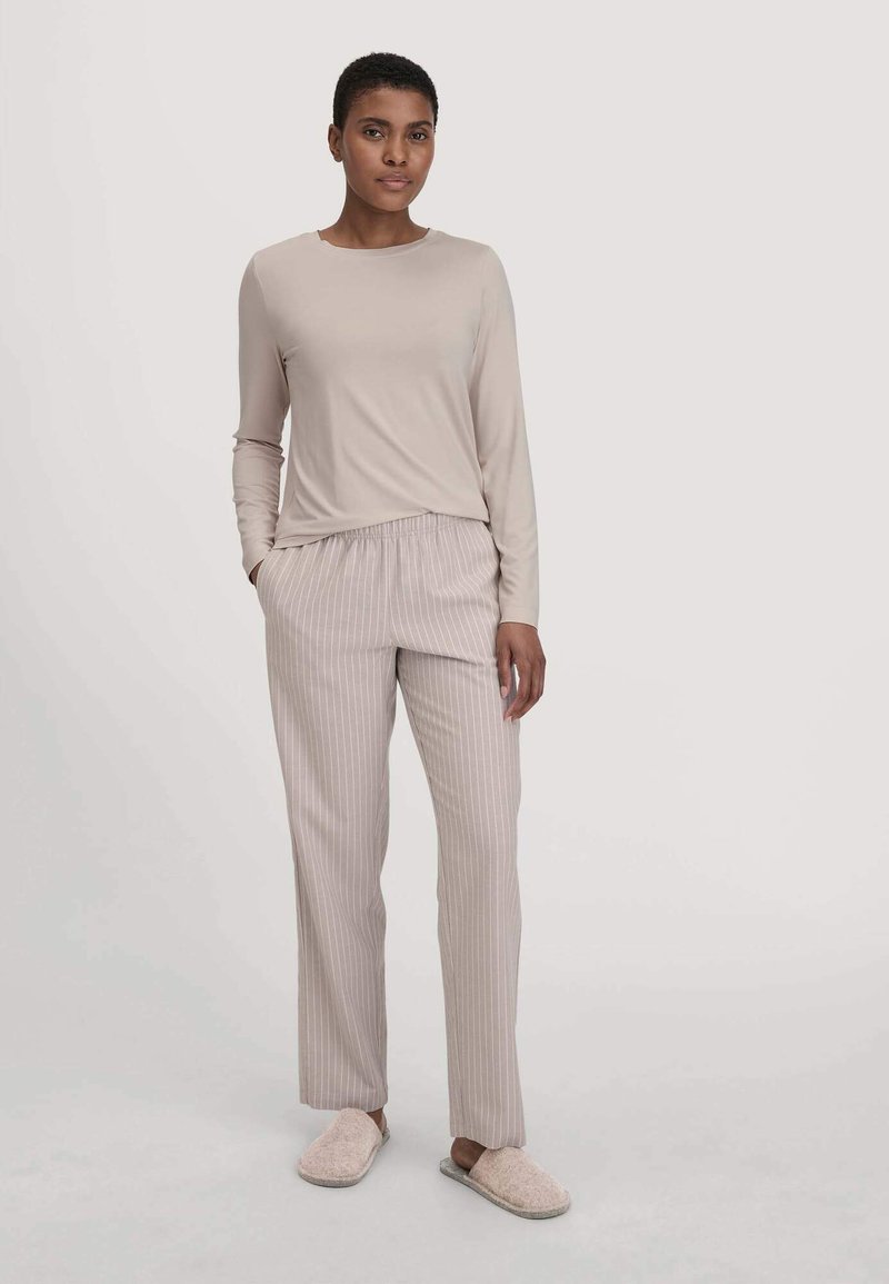 Frau steht mit einem beigen Langarmoberteil, hellbeigen, gestreiften Hosen und flauschigen beigen Hausschuhen vor einem einfarbigen Hintergrund.