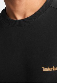 Felpa nera con colletto girocollo a coste e tessuto testurizzato; presenta un logo dorato "Timberland" sul davanti.