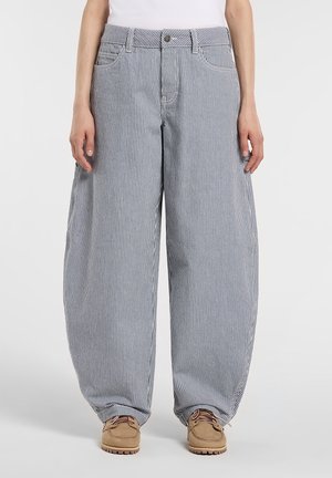 Personne portant un pantalon ample, taille haute, à rayures verticales bleues et blanches, associé à des chaussures à lacets couleur camel et un haut blanc.