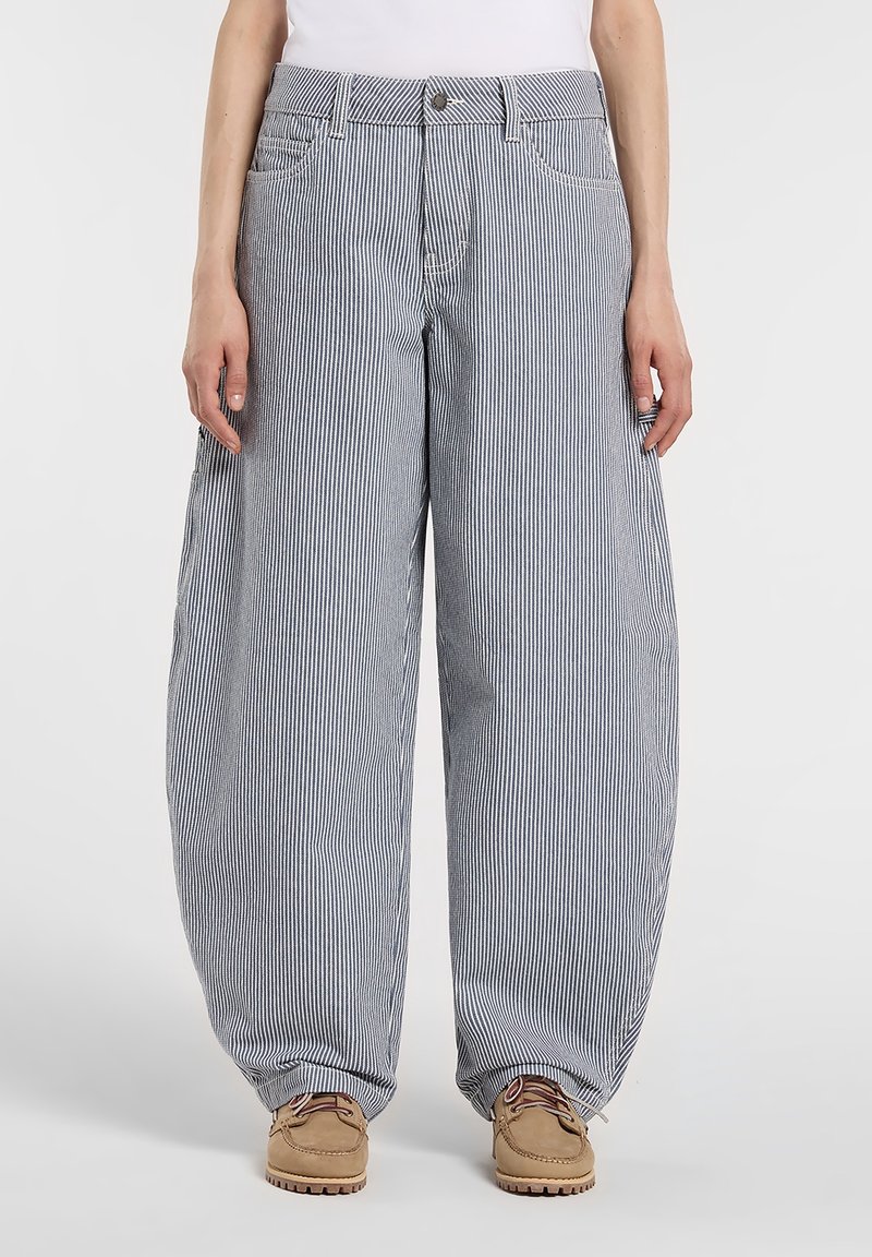 Personne portant un pantalon ample, taille haute, à rayures verticales bleues et blanches, associé à des chaussures à lacets couleur camel et un haut blanc.