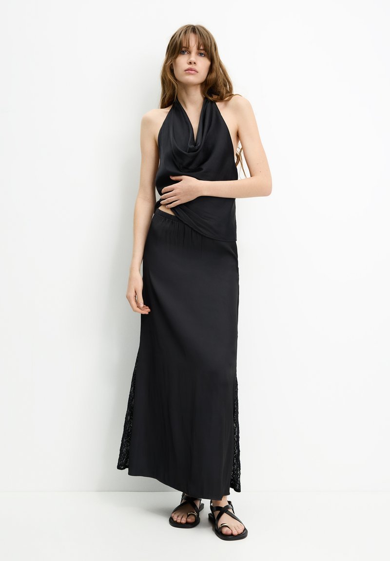 Schwarzes Halterkleid mit drapierter Brustpartie und langem Rock mit Spitzen-Seiteneinsätzen. Das Model trägt flache schwarze Sandalen. Glattes, leichtes Material.