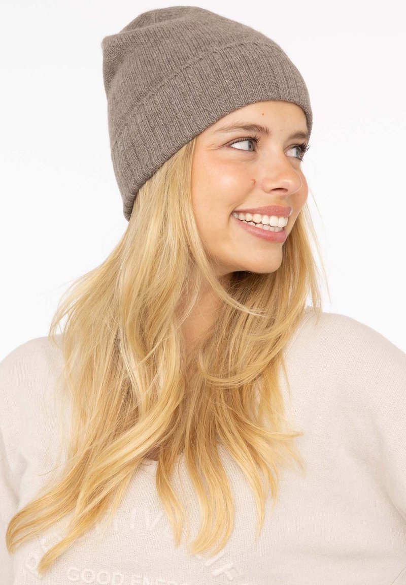 Brown knit beanie with a ribbed design, featuring a seamless top and a soft texture. Worn slightly slouched on a model with long hair.