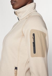 Veste zippée beige avec un motif texturé, col haut et écusson sur l'épaule. Comprend une poche zippée avec des accents foncés.