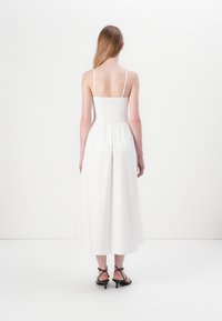 White, mid-length dress with thin straps and smocked bodice, featuring a flowing skirt. Worn with black strappy heeled sandals.