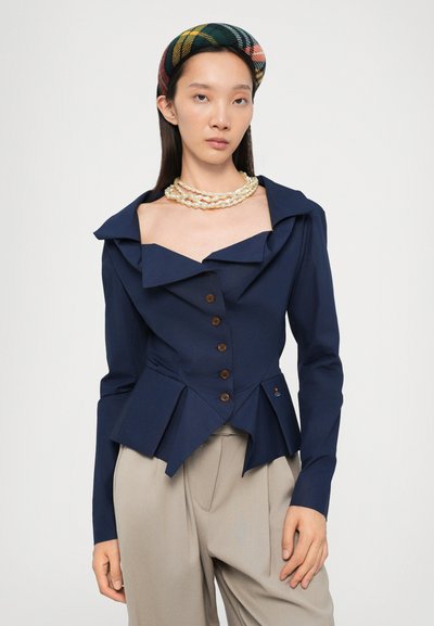 Navy blue collared jacket with large lapels, peplum waist, and wooden buttons. Model is wearing neutral trousers and layered necklaces.