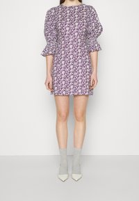 Robe à motif floral violet avec des manches bouffantes, longueur genou. Portée avec des chaussettes grises et des talons blancs pointus. Texture lisse, tissu léger.