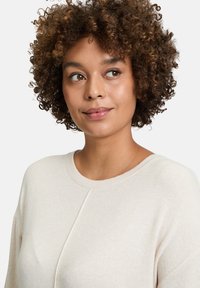 Cremfarbener Pullover mit rundem Halsausschnitt, der aus strukturiertem Stoff mit dezenten Nahtdetails gefertigt ist, getragen von einer Person mit lockigem Haar.